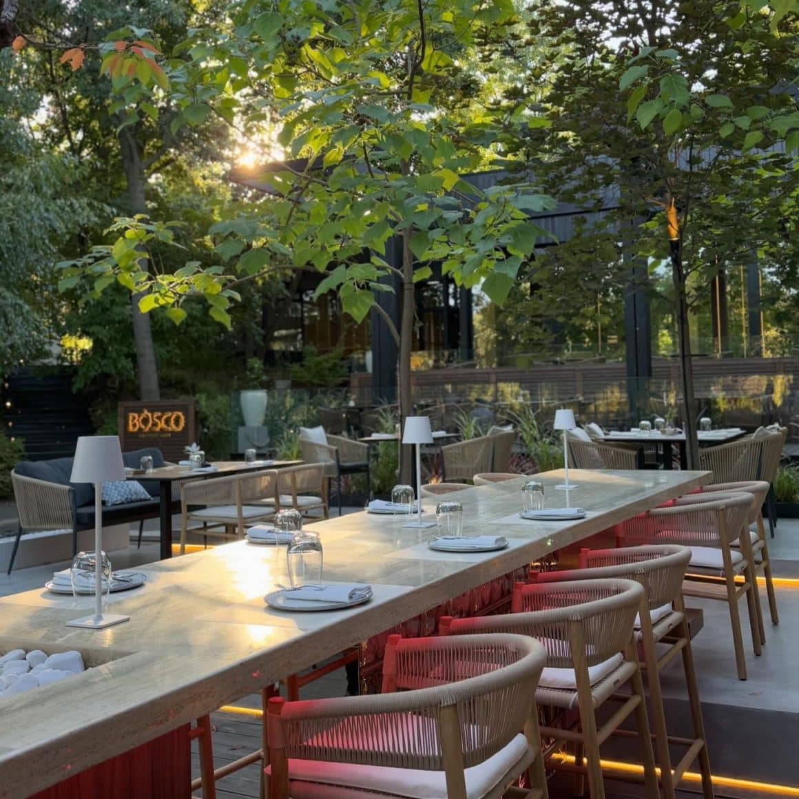Bosco Gourmet Nest București, cu masă comună în aer liber, mobilier de exterior și terasă înconjurată de vegetație.