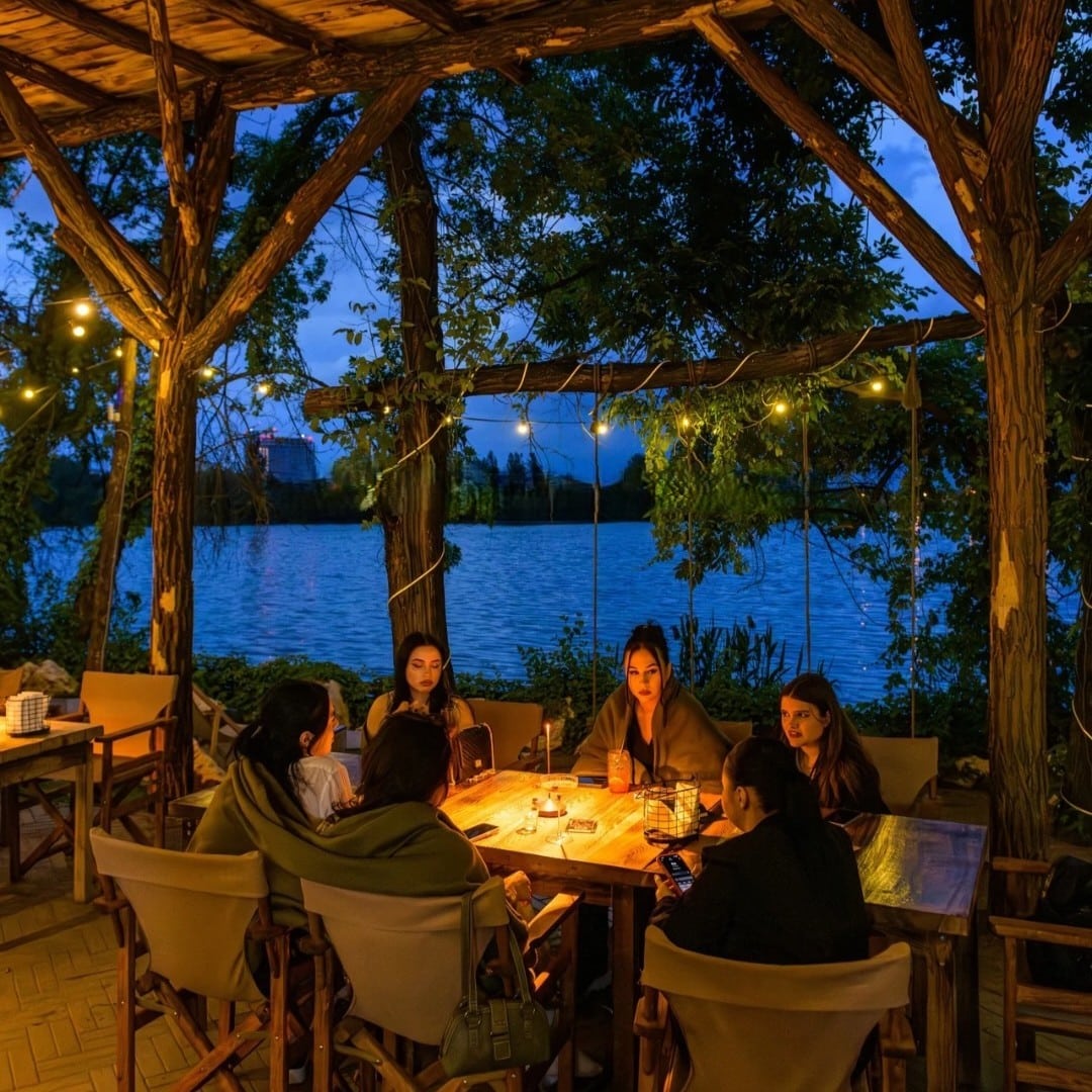 Seară de vară la terasa Indigen Space for Soul București – pergolă rustică din lemn, becuri și vedere la lac
