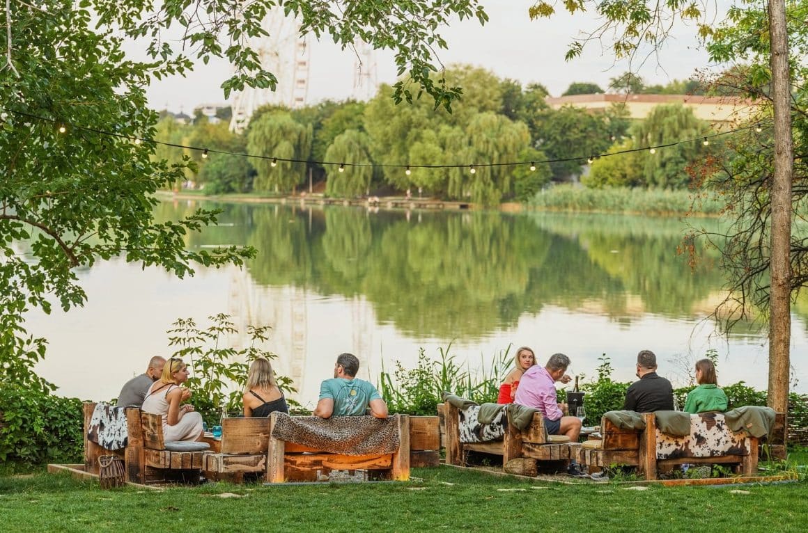 Terasă verde din București, pe malul lacului, cu mese rustice din lemn și oameni relaxați la apus. Terasa Indigen Space fort he Soul