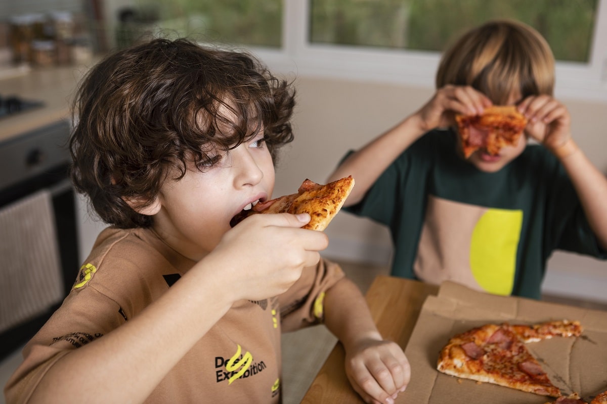Doi copii, baieti unul musca din felie de pizza, altul ridica o felie de pizza in dreptul fetei, ca si cum se ascunde