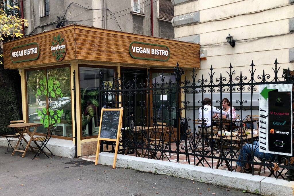 sara green victoriei, unul din restaurante vegetariene București, cu intrarea in terasa, imprejmuita cu gard din fier forjat si localul cu ferestre mari si stalpi de lemn pe care este trecuta firma