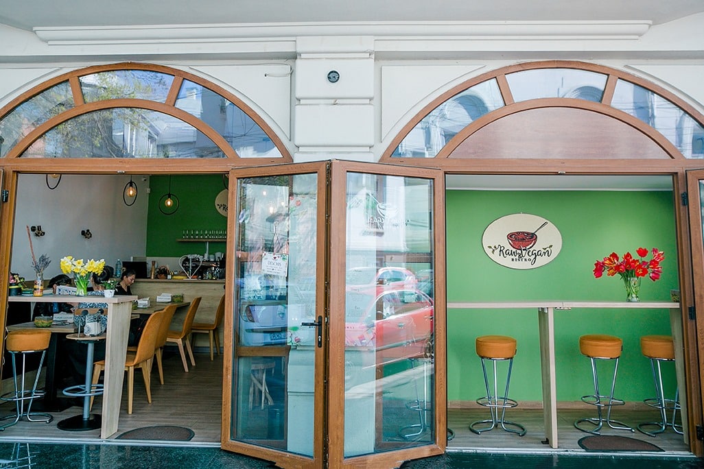 Vyro Raw Vegan fotografiat de afara, cu doua ferestre mari gen arcada, deschise larg, prin are se vede interiorul restaurantului, cu scaune inalte si pereti verzi