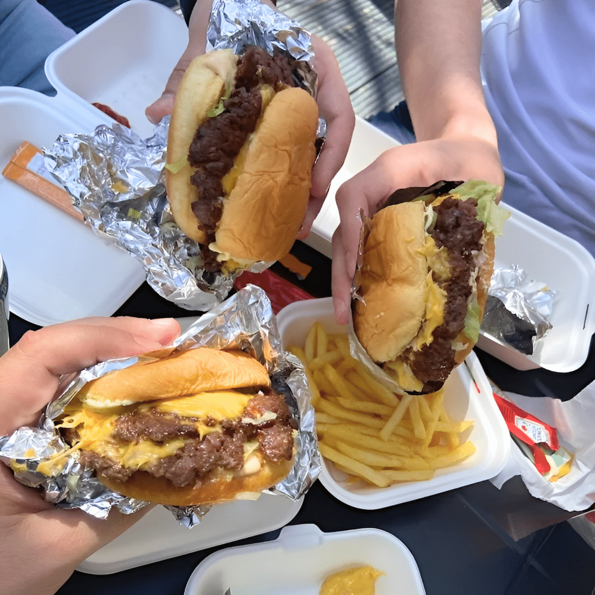 Trei burgeri Smash Baby desfăcuți din folie, cu carne smashed și brânză topită, lângă porții de cartofi prăjiți