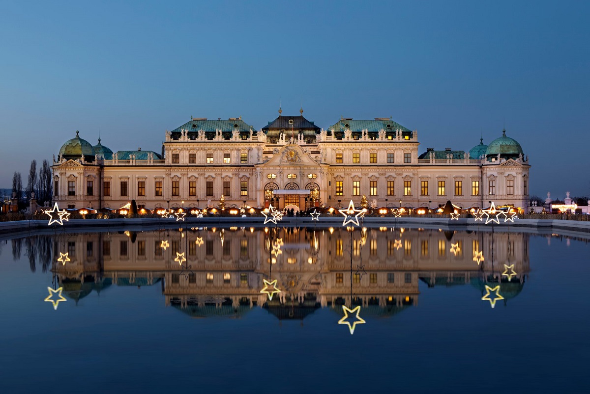 Targul de Craciun din fata Belvedere, Viena, cu un lac in prim plan iar in fundal cladirea impunatoare, istorica, iluminata