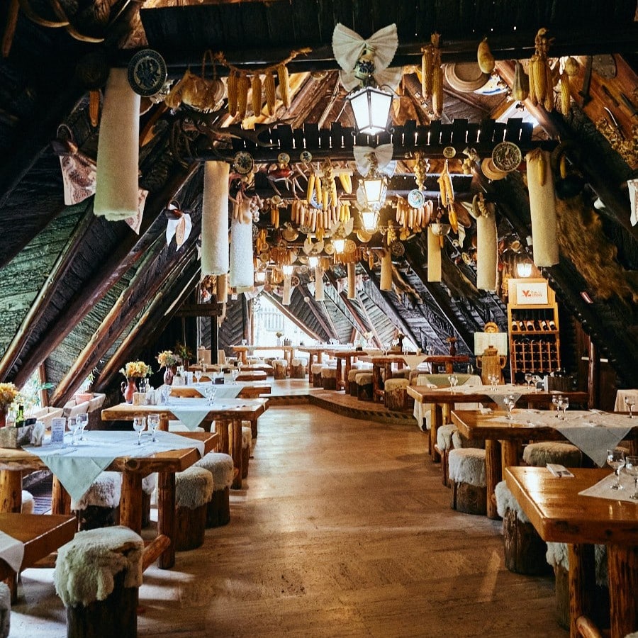 Restaurant rustic Șura Dacilor din Poiana Brasov, cu mese din lemn si tavan inclinat, din lemn, de care stau atarnate carnuri si alimente