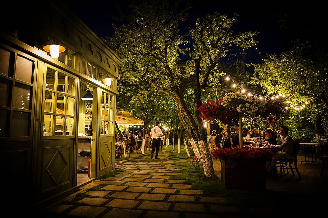Terasa de la restaurant Livada din Cluj, fotografiata noaptea, cu luiminite decorative, copaci si flori si oameni asezati la mese. restaurante Cluj
