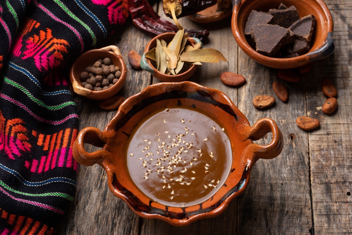 Mole, sos tradițional mexican, fotografiat de sus pe fundal de lemn, in vase d elut, alaturi d eboluri cu ciocolata neagra si aștul cu frunze de dafin, iar langa el o paturica cu motive mexicane