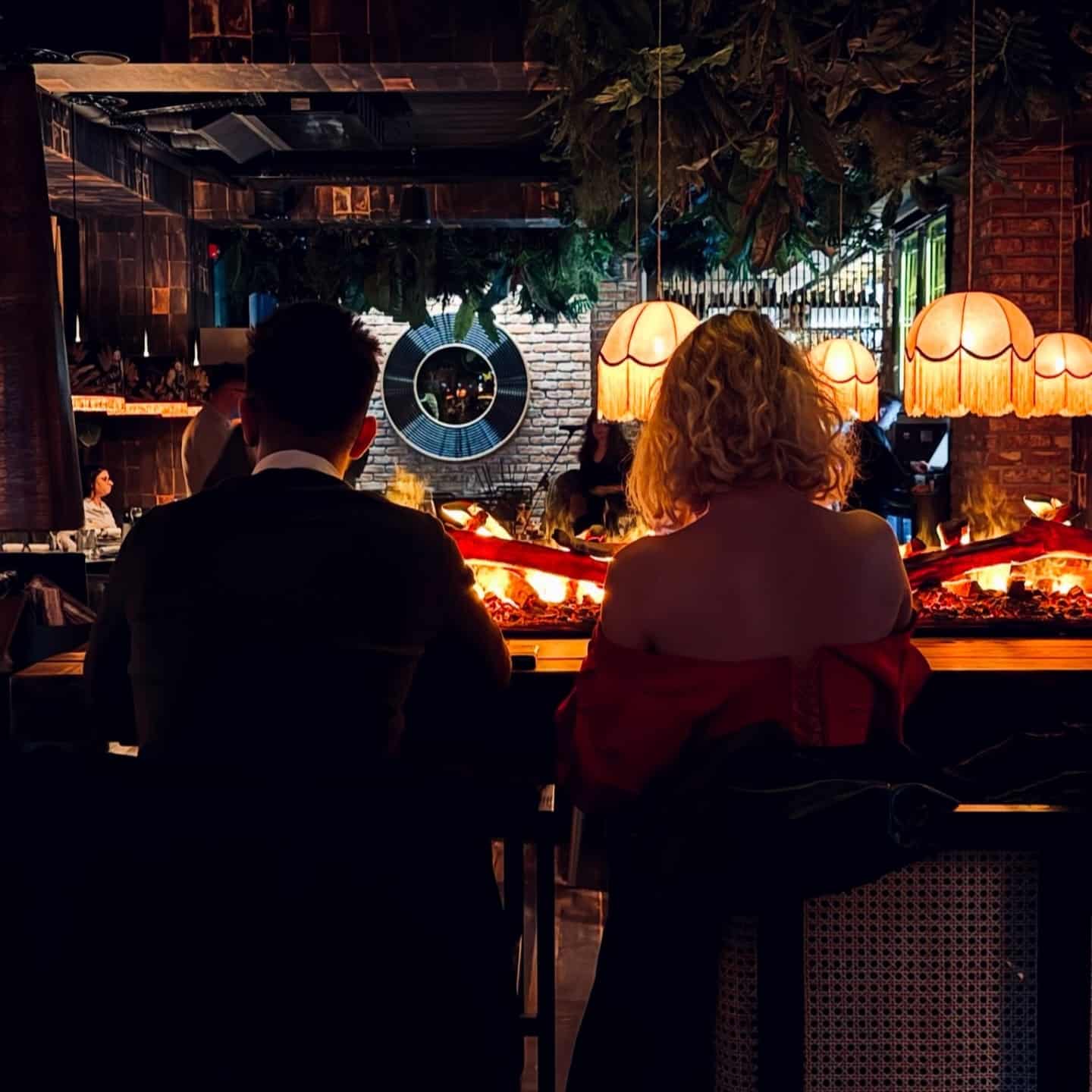 Cuplu la bar lângă șemineu deschis la Elephante București, cu lămpi cu franjuri și vegetație abundentă, atmosferă perfectă pentru o cină romantică de restaurante 8 martie