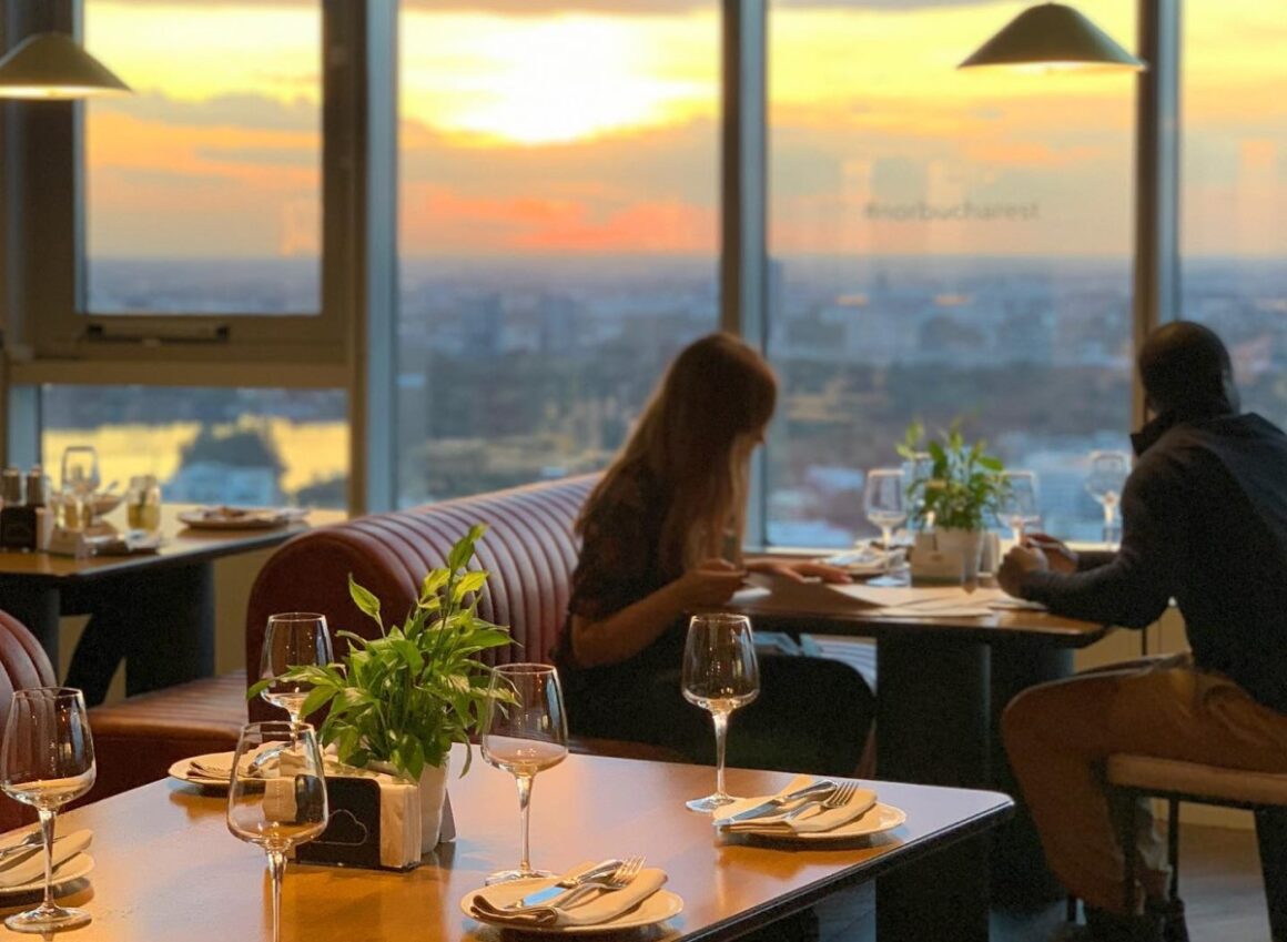  Cuplu care ia masa la restaurant in fata unor geamuri mari, la apus, cu vedere panoramica asupra orasului 