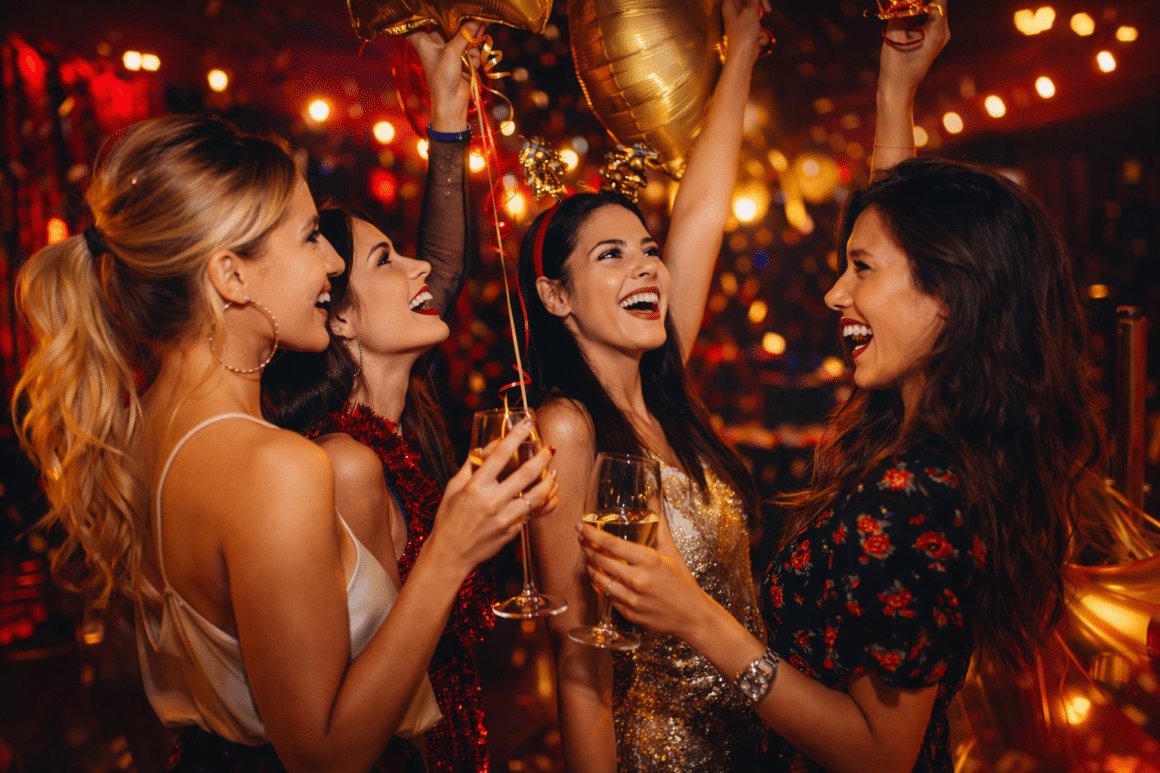 Patru prietene în ținute elegante dansează și râd la petrecere, cu pahare de șampanie și baloane aurii, lumini calde în fundal, atmosferă de girls night out de 8 martie