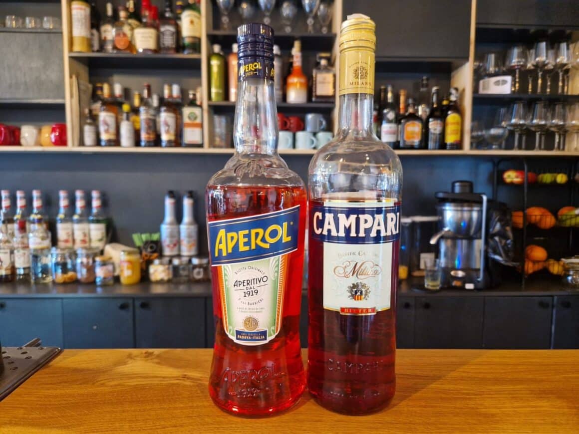 sticla de Aperol si Campari, asezate pe blatul unui bar, iar in fundal rafturi cu sticle
