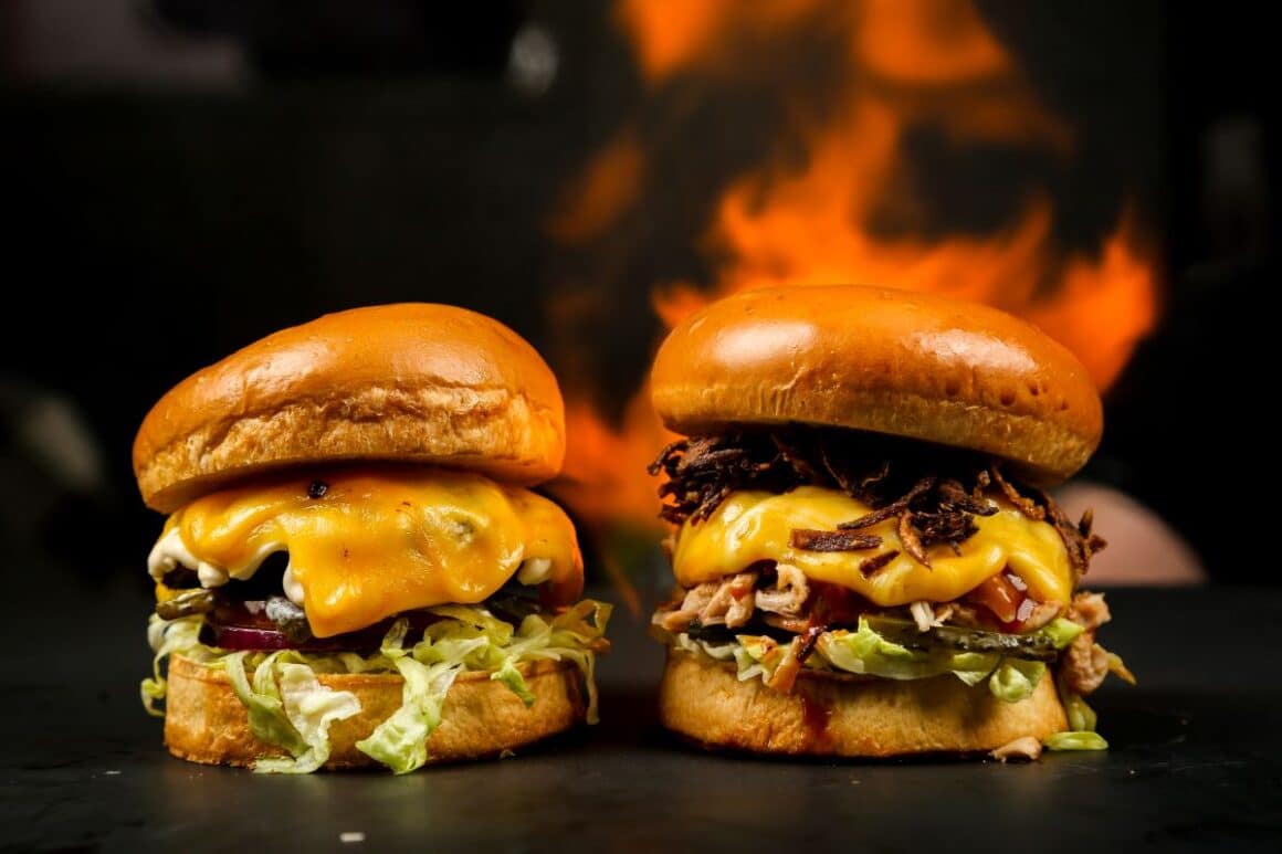 doi cheeseburgeri cu toppinguri diferite sunt asezati in fata unui foc 