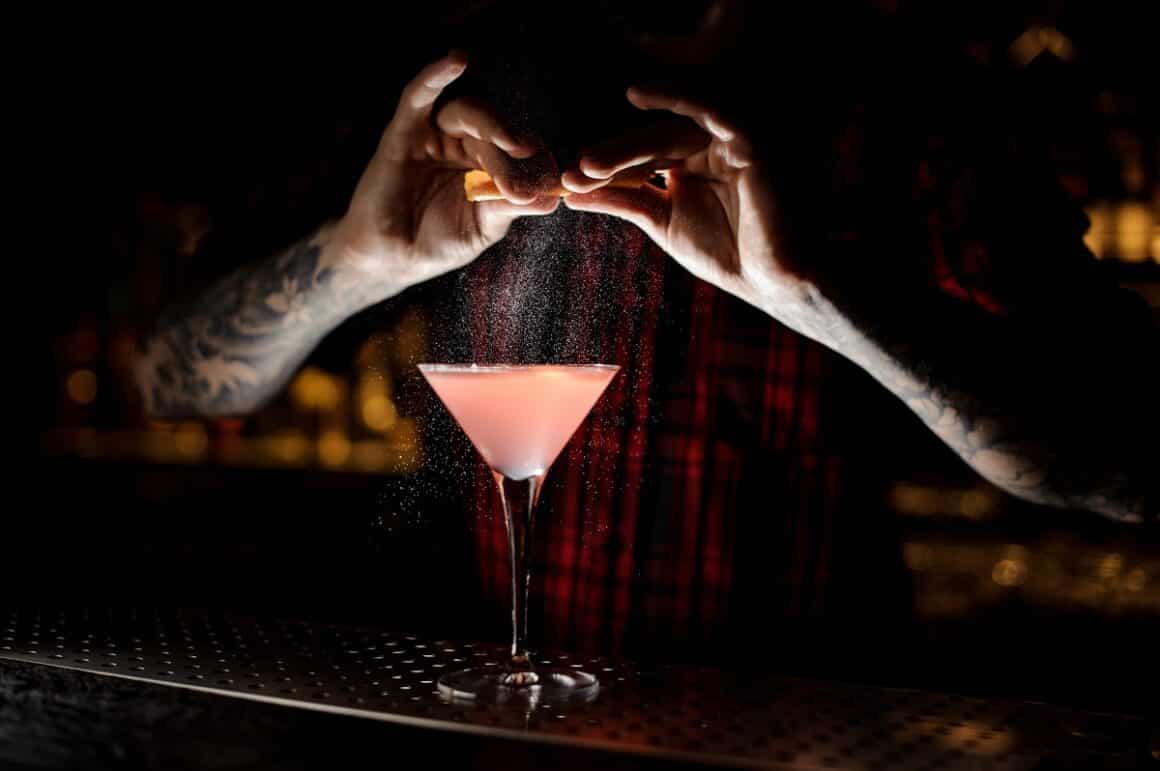 barman adauga zeama de portocala la un cocktail Cosmopolitan asezat pe bar, cu fundalul blurat