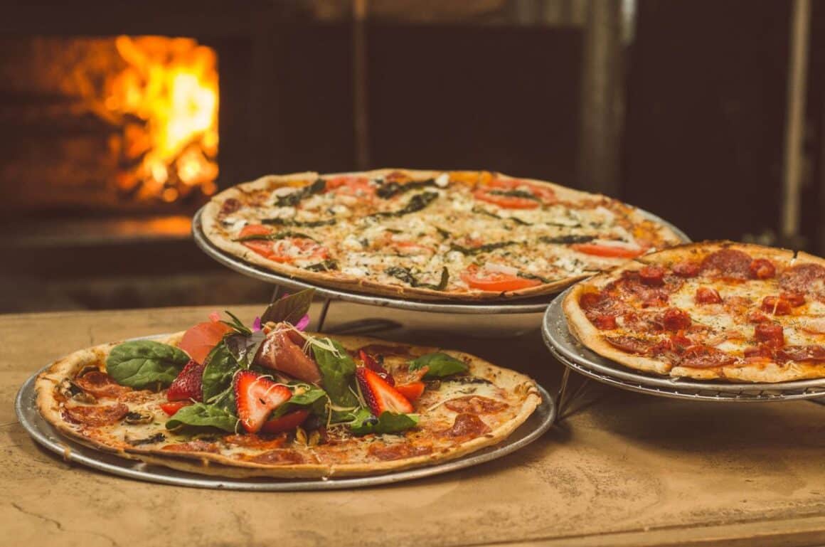 trei pizza sunt așezate in fata unui cuptor cu lemne traditional.