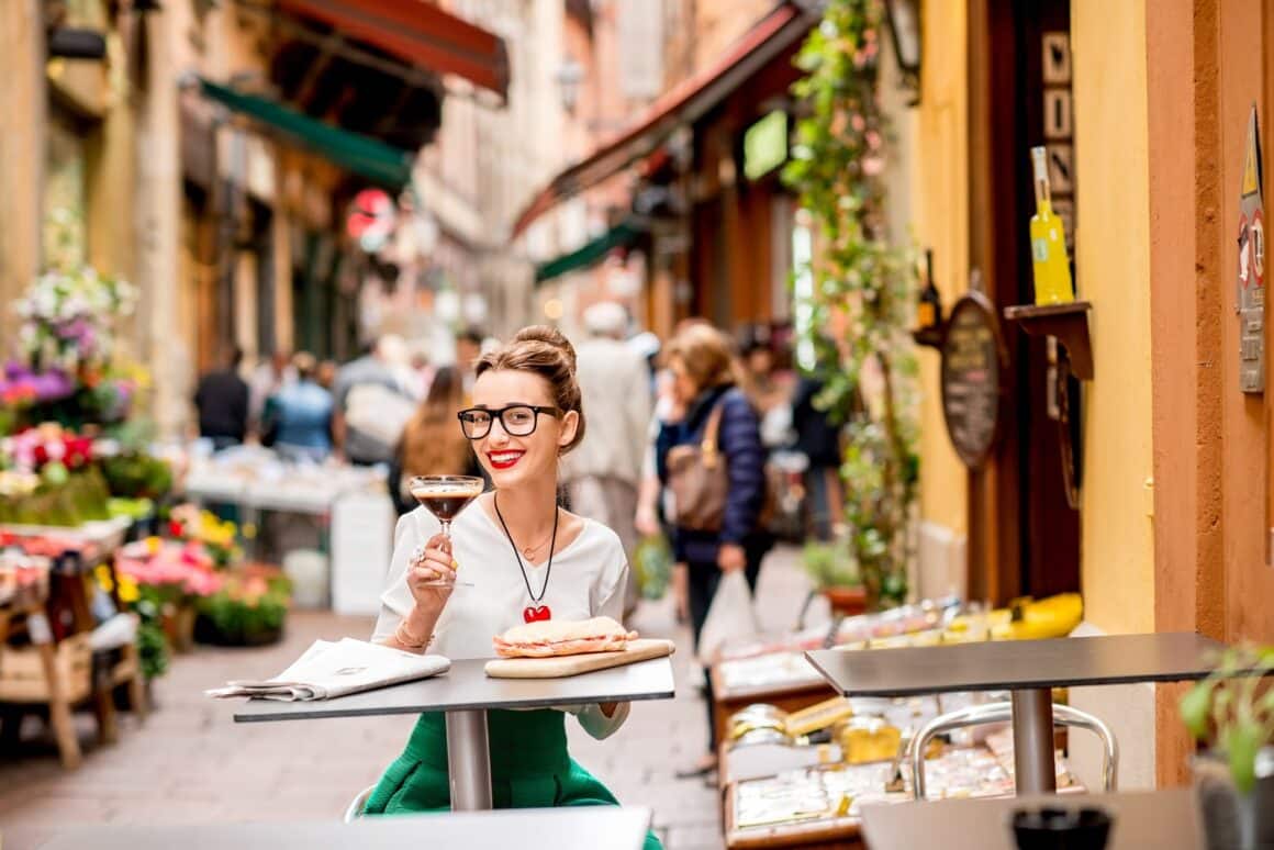 Young woman sitting at the cafe with shakerato coffee and panini outdoor on the famous street with local food markets in Bologna city.