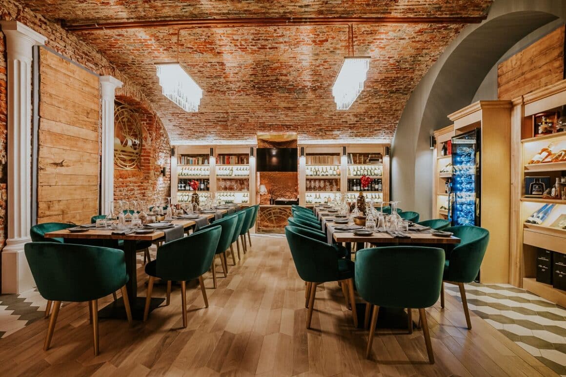 imagine din restaurant Allegria Oradea, cu pereti din caramida aparente, 2 mese lungi cu scaune capitonate verzi si in fundal rafturi cu dticle de vin