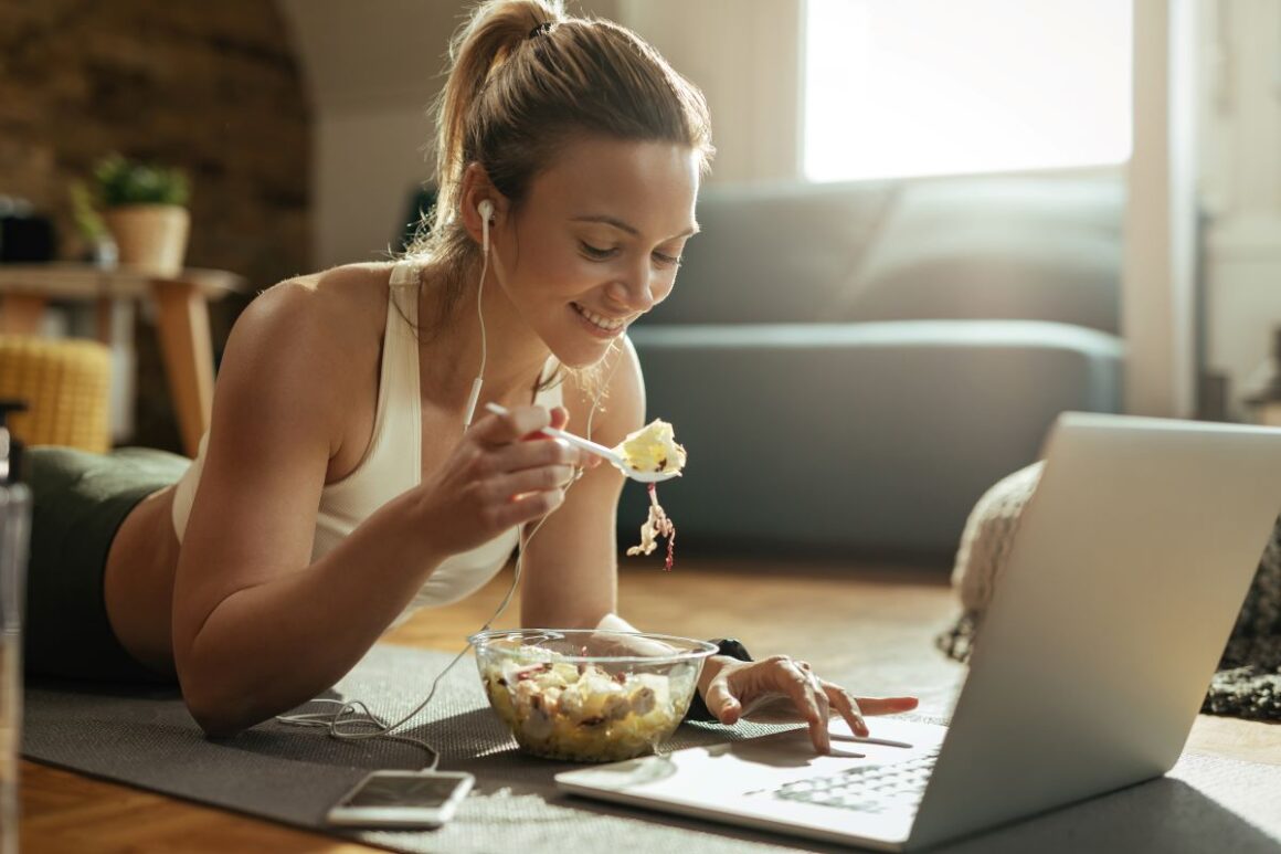o femeie în echipament de sport mananca o salata in timp ce priveste la laptopul ei, pe podeaua camerei - imagine concept pentru cele mai populare diete