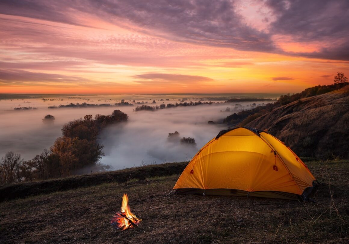 Cort asezat pe un deal, cu lumina aprinsa inauntru, un foc mic langa si in fundal valea acoperita de ceata si cerul la apus. Campinguri din România.