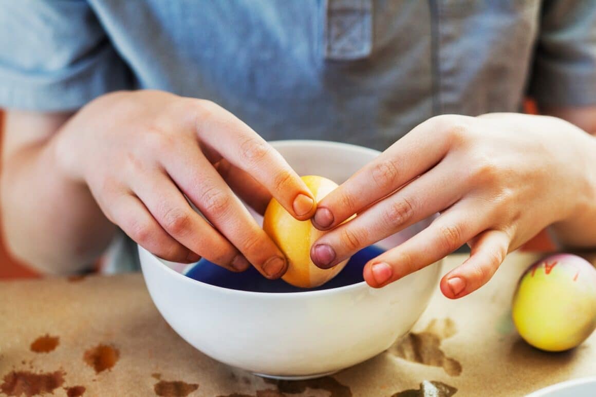 close up cu mainile unui copil care tine un ou galben deasupra unui bol alb cu vopsea