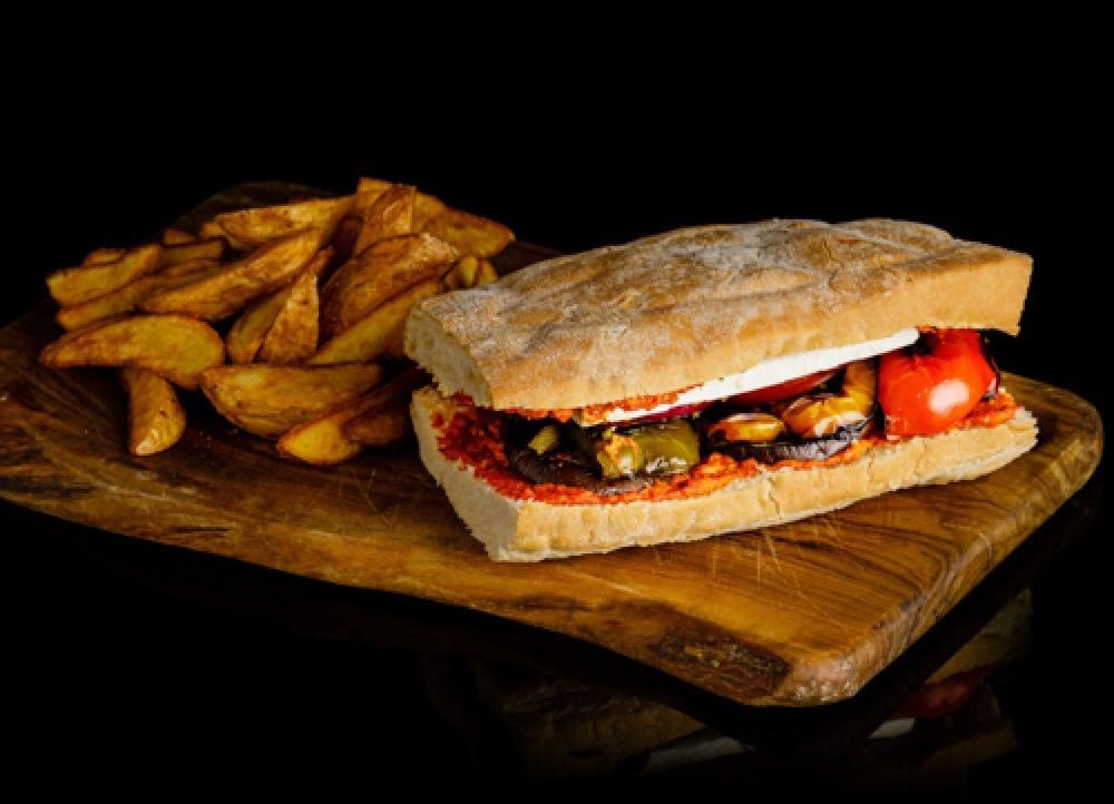 sandwich vegan panini servit pe un tocator de lemn, alaturi de cartofi prajiti, fotografiat pe fundal negru de la Pio Bistro 