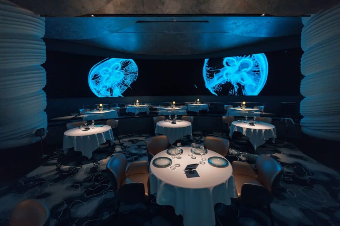 interiormese rotunde in restaurant ossiano dubai, cu lumina difuza si un perete negru pe care sunt pictate meduze florescente