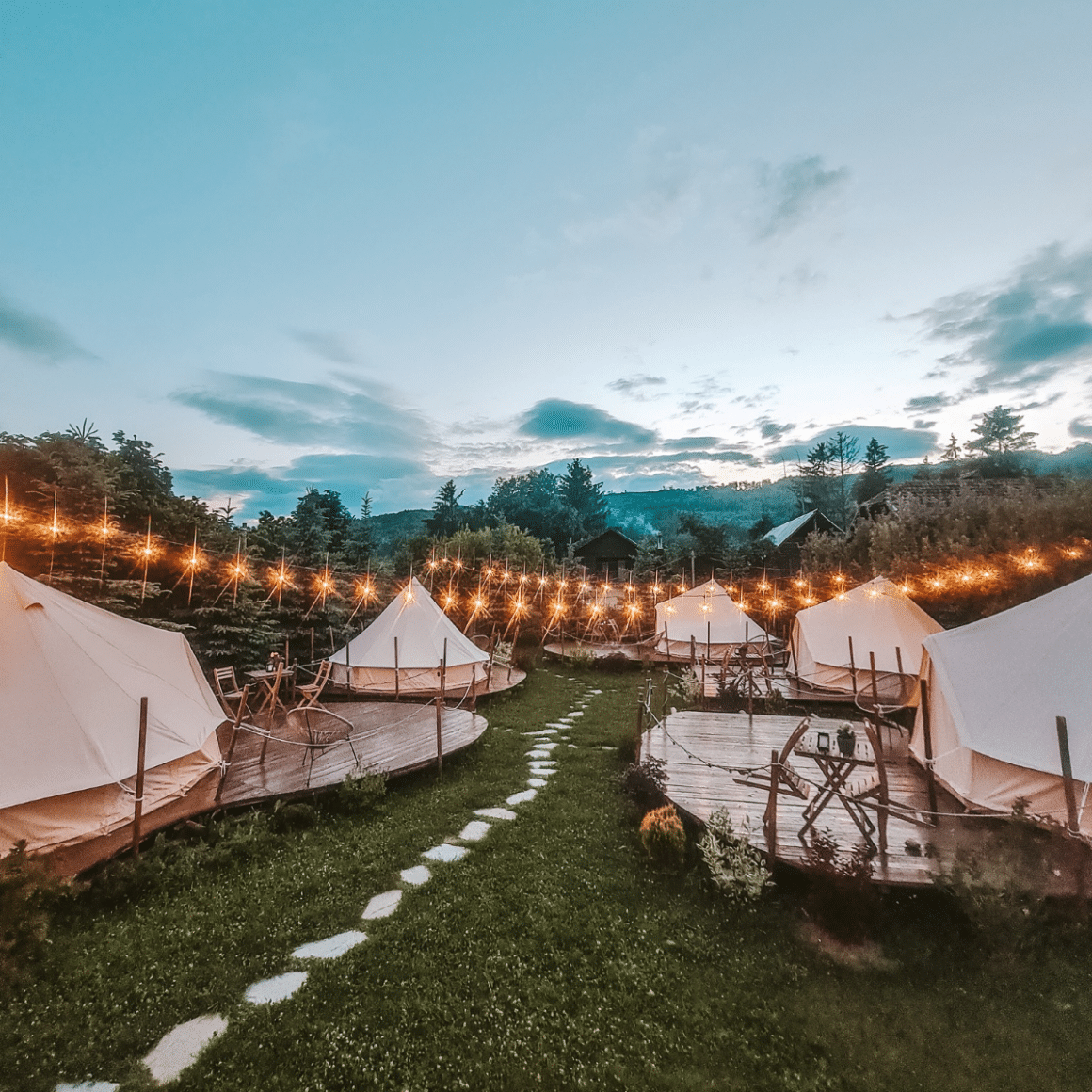 Complexul de glamping în România Valea Doftanei Glamping, cu mai multe corturi asezate pe gazon si marginite de copaci, cu luminite aprinse printre ele