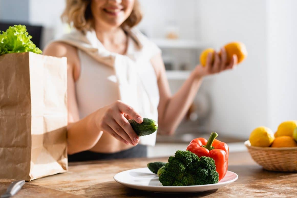 Poza cropp cu o femeie care aseaza un castravete pe o farfurie langa broccoli si ardei gras roșu, alimente pentru piele frumoasă și sănătoasă