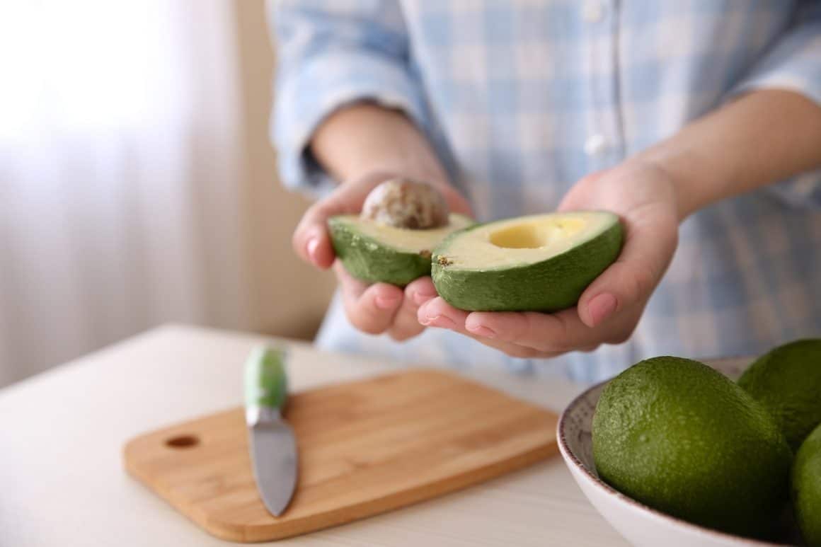 Close up cu mainile unei femei care tine un avocado taiat pe jumatate, unul din alimente pentru piele frumoasă și sănătoasă