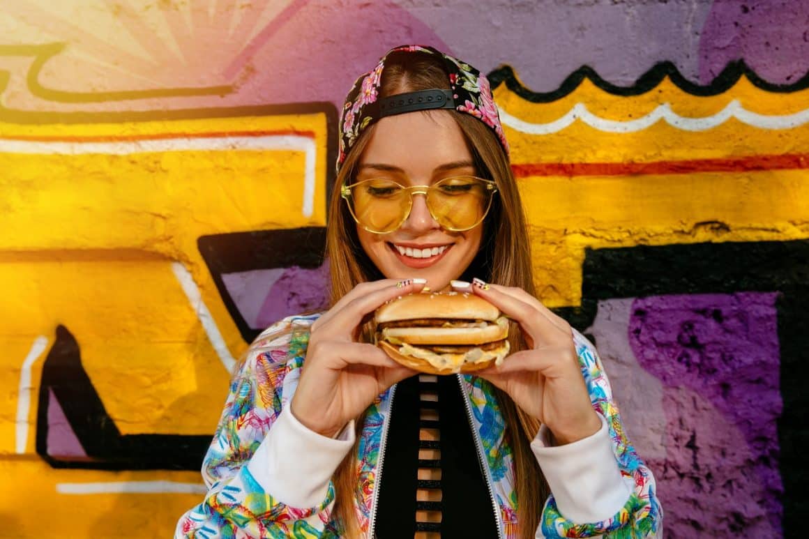 Femeie tanara zambeste int imp ce tine in mana un burger. E imbarcata colorat, cu sapca si ochelari, iar in spate e un perete cu grafitti. 10 burgeri gustoși în București