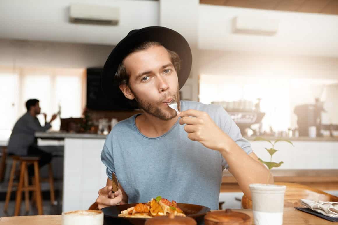 Fashionable young man enjoying tasty food for lunch sitting at wooden table of cozy restaurant. Hungry hipster wearing trendy black hat stilling his hunger while having meal at cafeteria alone