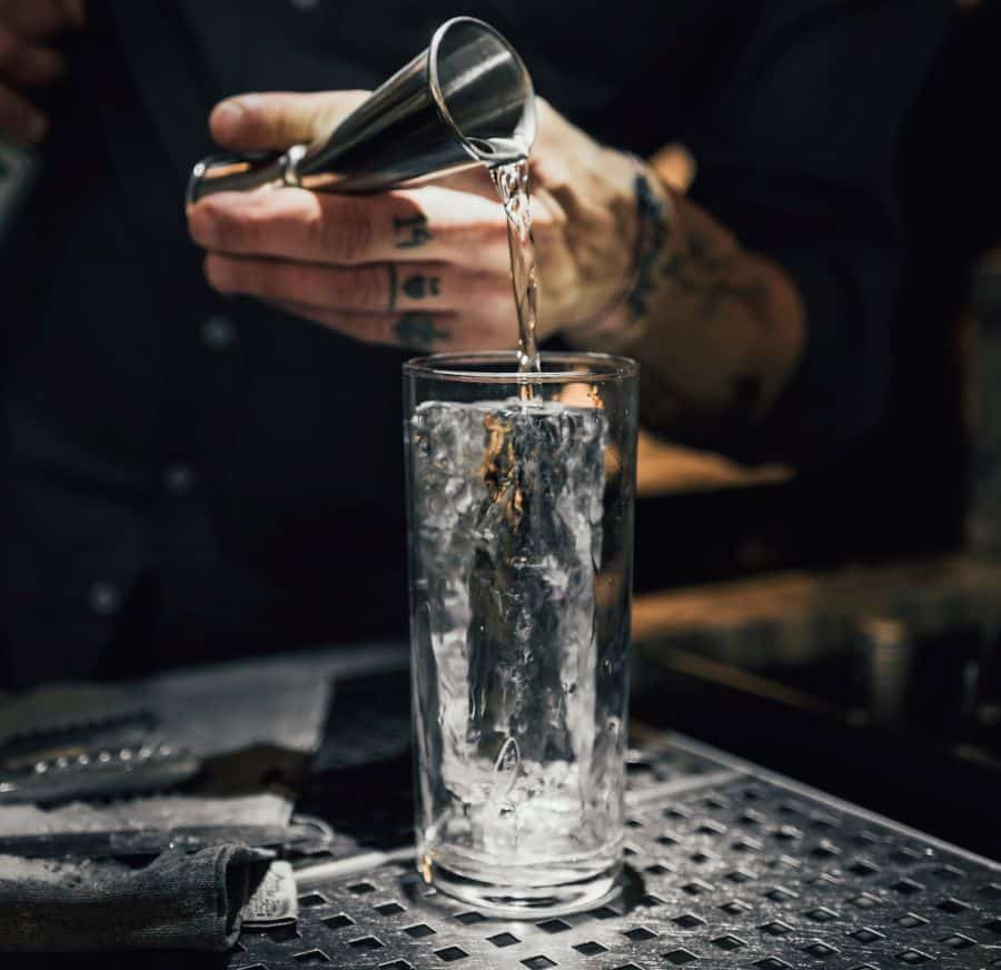 un barman toarna o masura de alcool peste doua cuburi de gheata intr-un pahar highball. Imagine pentru forma paharului de cocktail highball