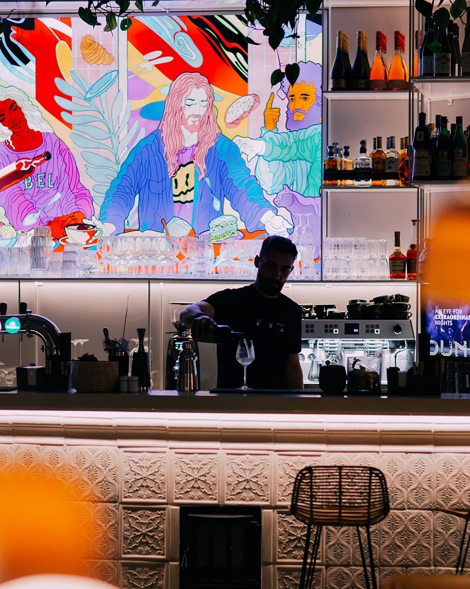 barman fotografiat in penumbra la The Other Saint, iar in spate un mural cu scena biblica interpretata modern. Restaurante bune pe Calea Victoriei