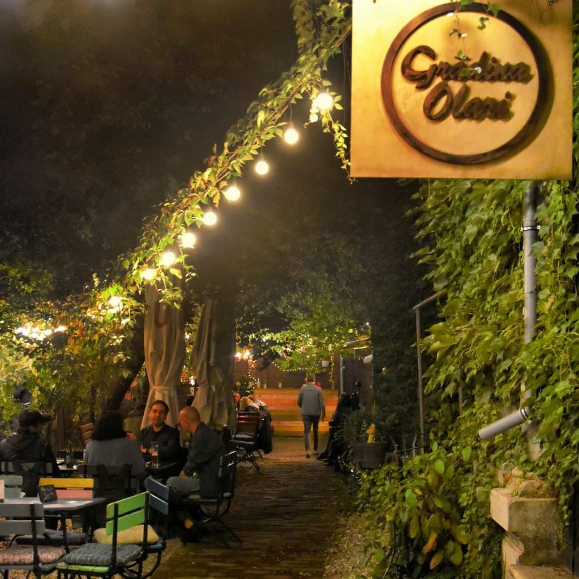 Gradina Olari fotografiata seara, plina de verdeata, cu luminite decorative si oameni care iau masa