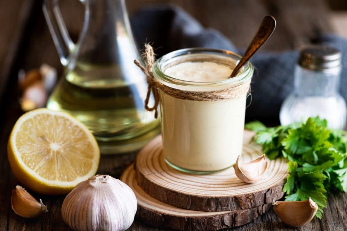 Un borcan de sos alioli este asezzat pe două rondele de lemn, alături de ingredientele necesare: lamaie, verdeață, sare, usturoi si ulei de masline