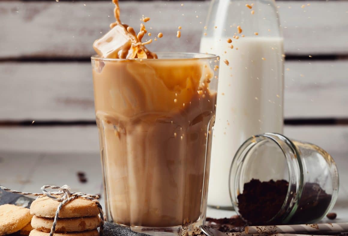 un pahar de ice latte din care sar cateva picături este asezatpe o masă alaturi de o sticlă de lapte, cativa biscuiti și un borcan cu pudră de cacao