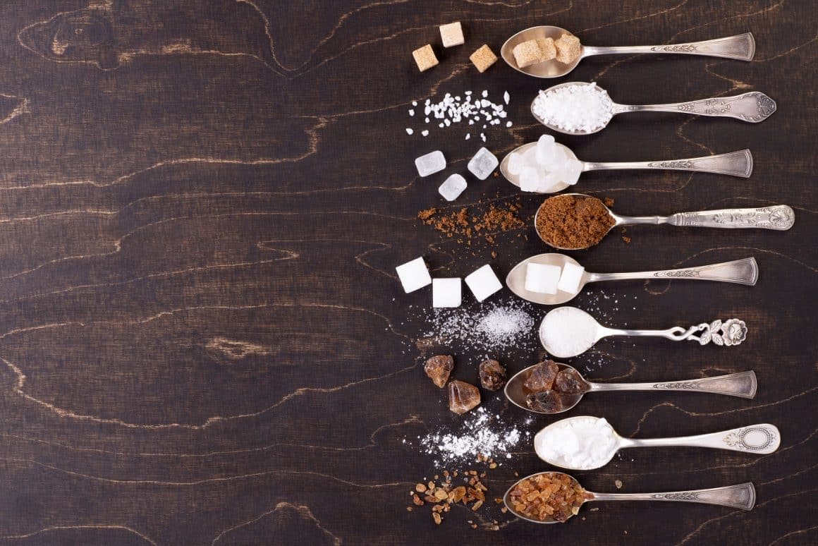 Mai multe lingurite aliniate, toate pline cu diferite tipuri de zahăr, fotografiate pe fundal de lemn