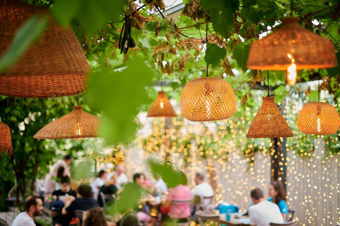 mai multe abajururi din rachită atarna deasupra unei grădini din București, cu mai multe mese si scaune cu oameni și ghirlande luminoase in fundal. La Gspot Sufrageria.