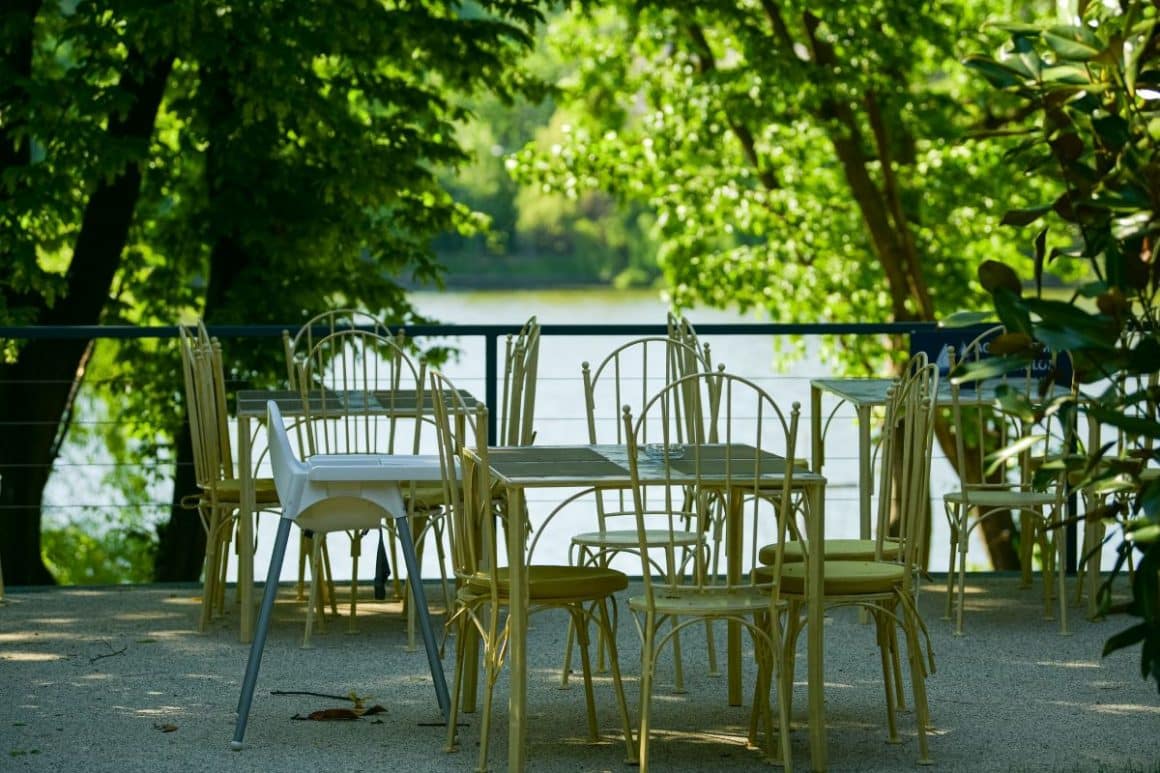 mai multe scaune sunt asezate in jurul unei mese din fier forjat pe terasa restaurantuluizexe, iar in fundal se vede lacul înconjurat de verdeată. Terase din Herăstrău