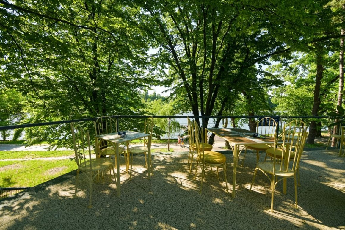 mai multe scaune sunt asezate in jurul unei mese din fier forjat pe terasa restaurantuluizexe, iar in fundal se vede lacul înconjurat de verdeată