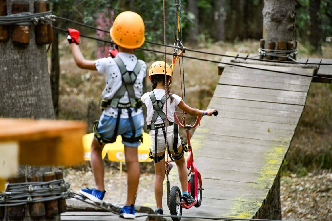 doi copii echipati de catarari, unul merge pe langa bicicleta lui pe un podet de lemn, suspendat intre copaci, la Neptun Paradis Land. Destinații de vară pentru familiile cu copii