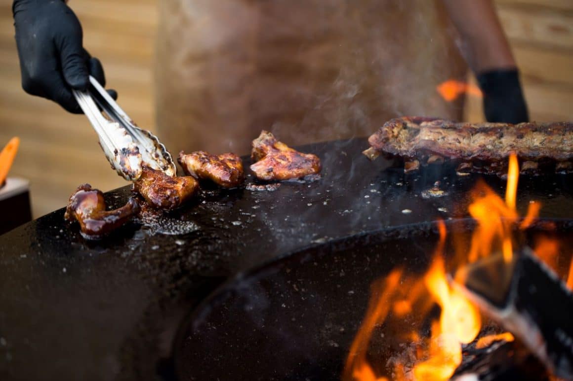 un bucătar prăjește pe marginea unei plite rotunde mai multe bucăți de aripioare picante, iar în prim plan se văd câteva limbi de foc