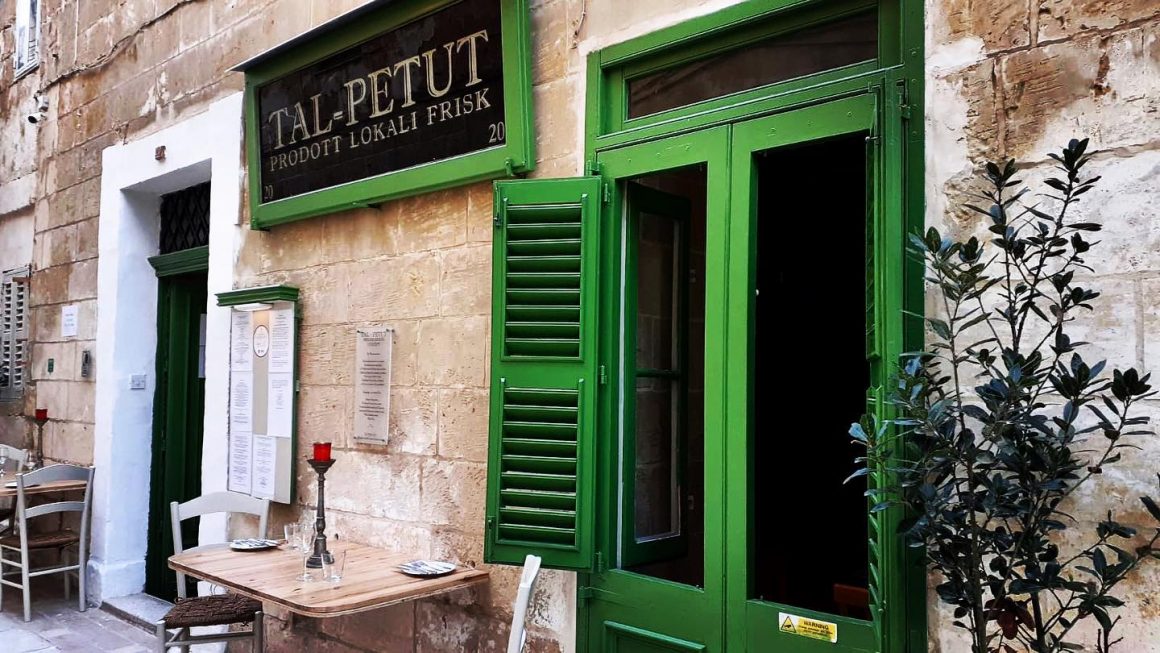 intrarea in restaurantul maltez Tal-Petut cu geamuri si obloane verzi și o masă de lemn cu doua scaune