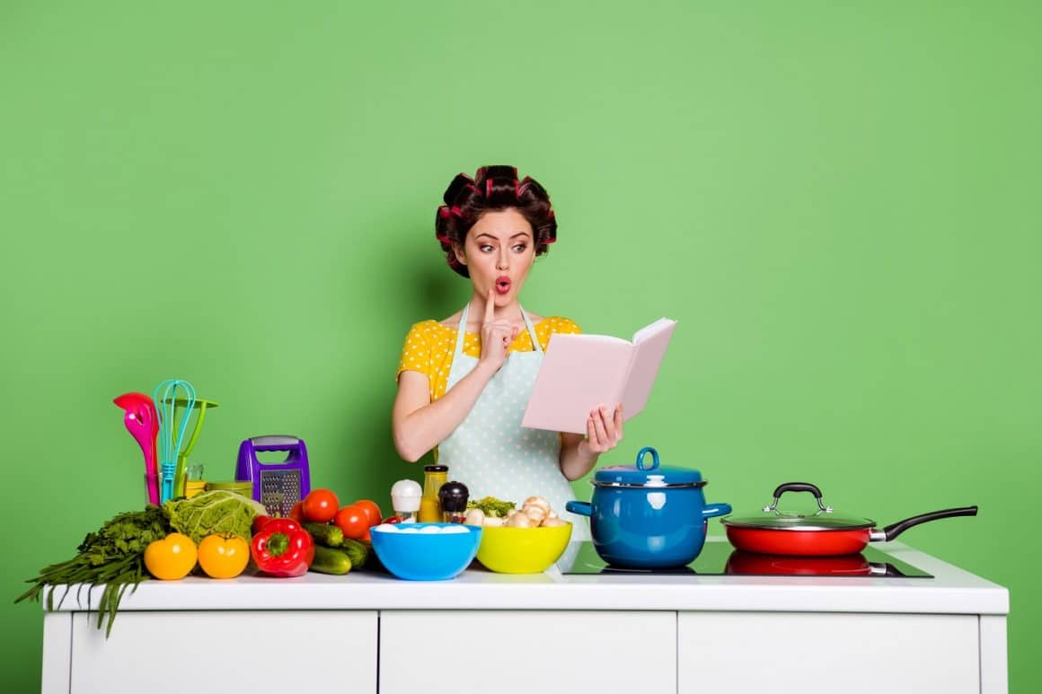 Femeie cu sort de bucatarie si bigudiuri citeste o carte de bucate in fata unui blat d ebucatarie plin cu fructe, legume, si oale colorate. Imagine reprezntativă pentru reguli  la gătit.