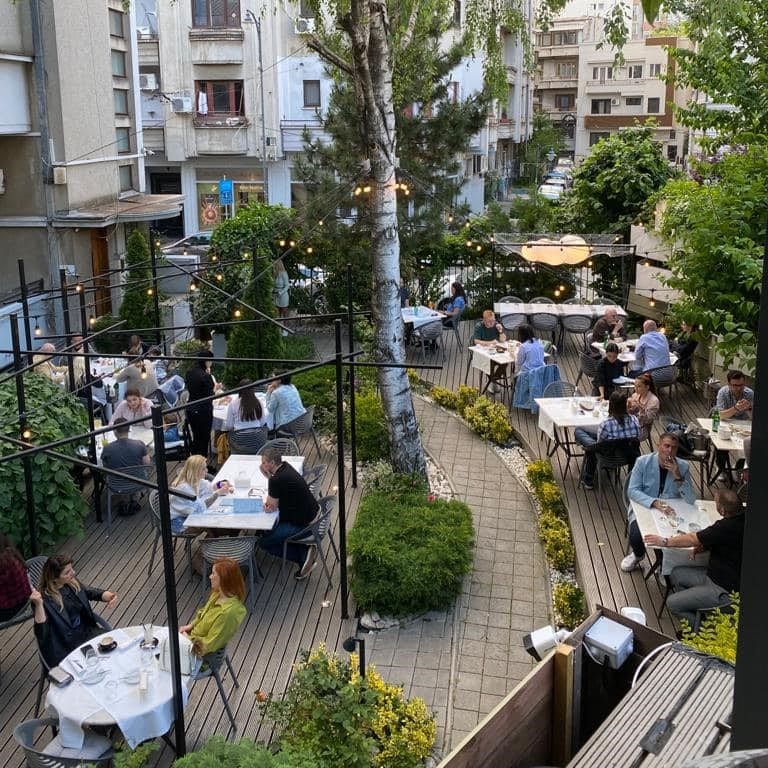 Mai multe mese la care oameni iau masa pe terasă în grădina restaurantului savart București
