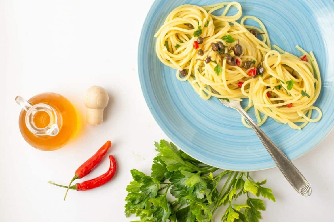 Farfurie cu paste Spaghetti alla colatura, in farfurie albastra, fotografiata de sus, pe fundal alb alaturi de patrunjel proaspat, ardei iute si sticluta cu ulei. Preparate tradiționale din Amalfi
