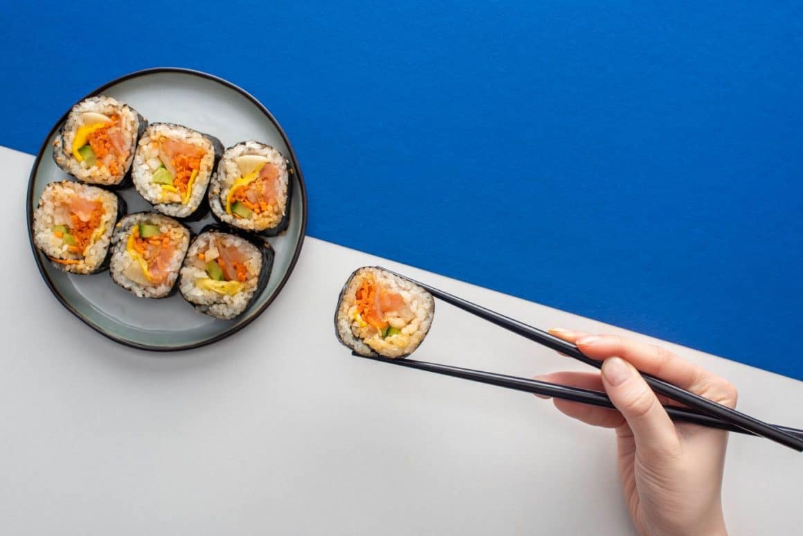 o mana de femeie tanara apuca cu ajutorul betisoarelor chinezesti un rulou de sushi de pe un platou asezat pe o suprafata jumatate alba jumatate albastra. Cum se mănâncă sushi