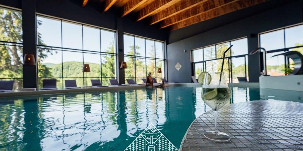 un pahar de cocktail este fotofrafiat pe marginea piscinei interioare a resortului balvanyos, iar in plan secund, afara se vad brazii