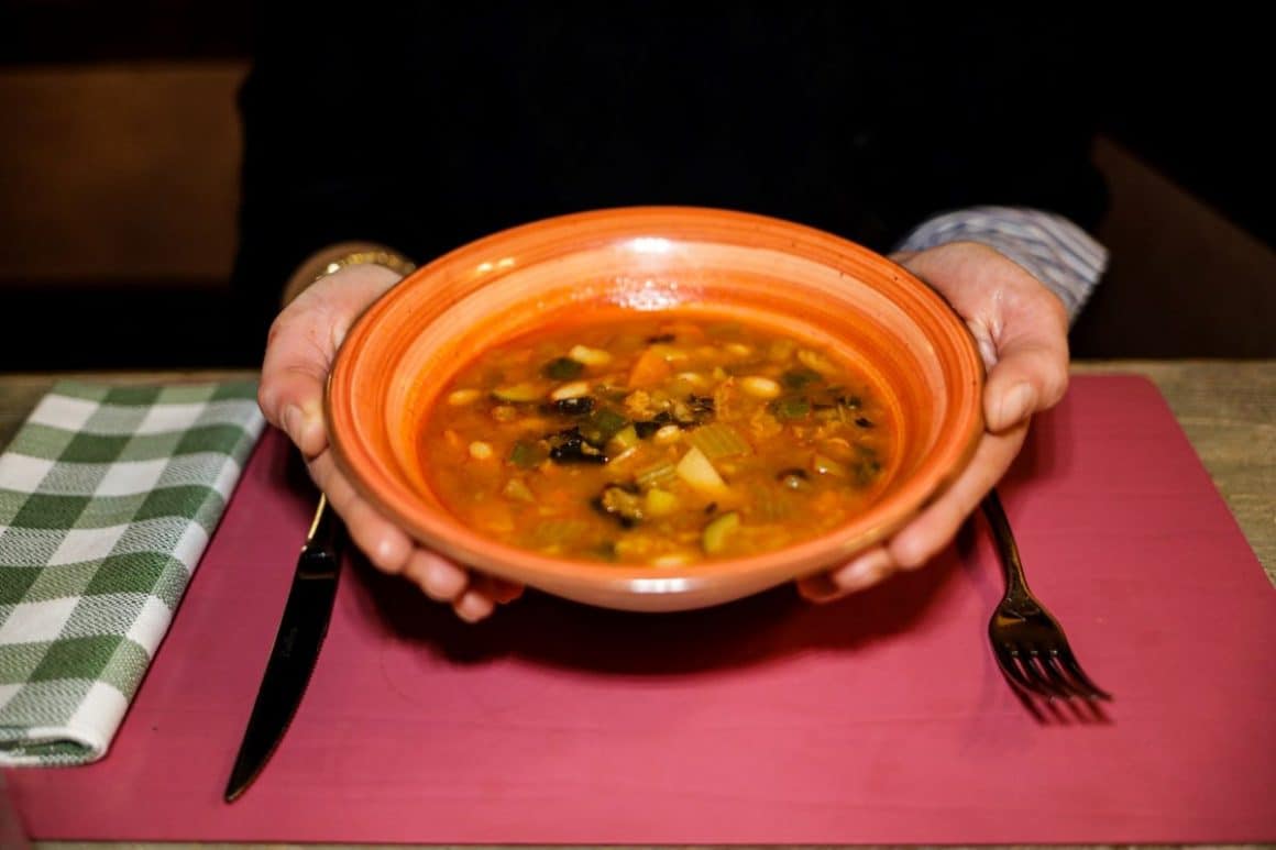un barbat tine cu ambele maini deasupra unei mese un bol cu Supa ribollita