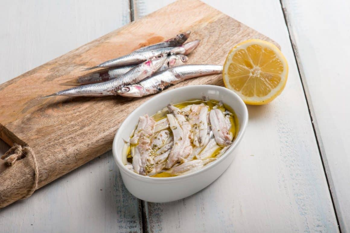 pe un tocător de bucătărie sunt asezati cativa pești, iar langa, pe aceeași masă, este un bol cu file de ansoa marinate în ulei, cu o jumatete de lamaie langa