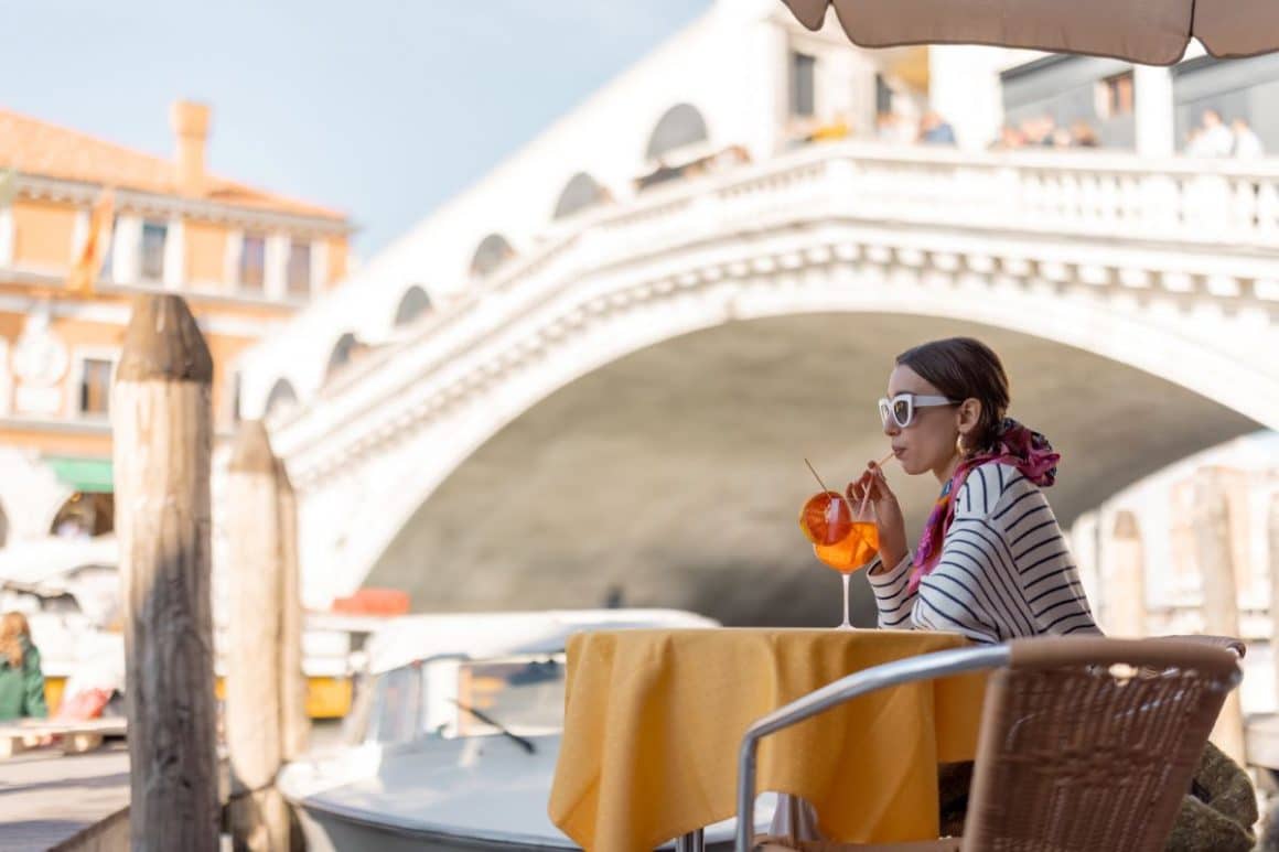 o femeie bea cu paiul dntr-un pahar de aperol sprit, la una dintre terasele de langa un pod venetian